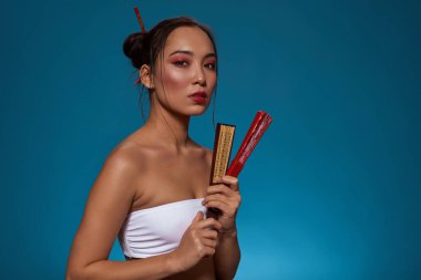 Serious elegant asian girl holding two closed traditional handheld fans and looking at camera. Beautiful young brunette woman wearing tank top. Female beauty. Blue background. Studio shoot. Copy space