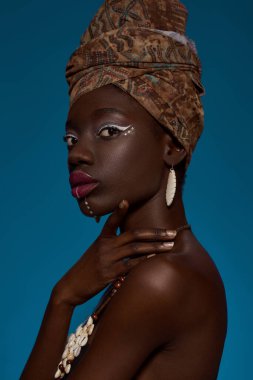 Cropped side view of fashionable black girl wearing traditional african outfit and accessories. Pretty young slim woman looking at camera. Female beauty. Isolated on blue background. Studio shoot