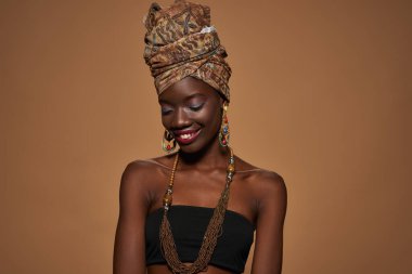 Smiling elegant black girl wearing traditional african outfit and accessories. Beautiful young slim woman wearing tank top, turban, necklace and earrings. Orange background. Studio shoot. Copy space