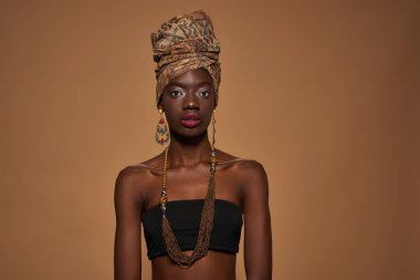 Front view of elegant black girl wearing traditional african outfit and accessories looking at camera. Young woman wear tank top, turban, necklace and earrings. Orange background in studio. Copy space