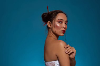 Side view of elegant korean or chinese girl looking at camera. Beautiful young brunette slim woman wearing tank top. Female beauty. Isolated on blue background. Studio shoot. Copy space