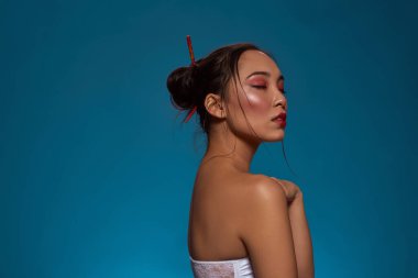 Side view of stylish japanese and chinese girl with closed eyes. Attractive young brunette slim woman wearing tank top. Female beauty. Isolated on blue background. Studio shoot. Copy space