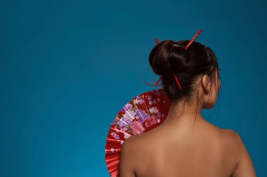 Back view of elegant asian girl with traditional handheld fan. Cropped of beautiful young brunette slim woman. Female beauty. Isolated on blue background. Studio shoot. Copy space
