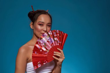 Confident elegant asian girl posing with traditional handheld fan and looking at camera. Beautiful young brunette slim woman wearing tank top. Female beauty. Blue background. Studio shoot. Copy space