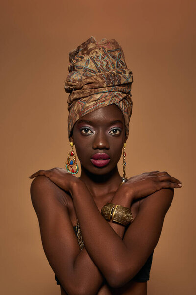 Elegant black girl wearing traditional african outfit and accessories looking at camera. Beautiful young woman wear tank top, turban, necklace, earrings and bracelet. Orange background. Studio shoot