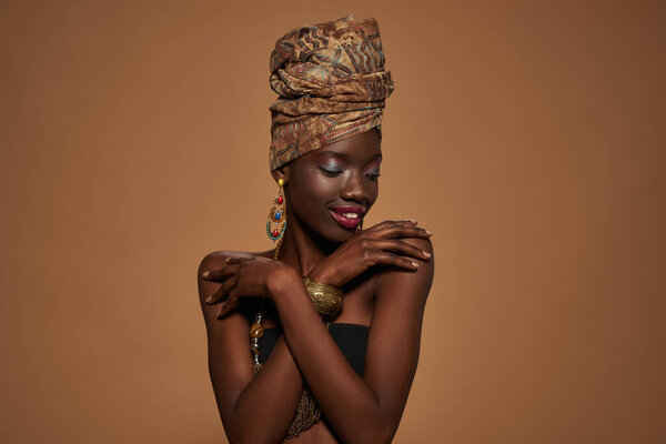 Smiling fashionable black girl wear african outfit and accessories hug herself. Young woman wearing tank top, turban, necklace, earrings and bracelet. Orange background. Studio shoot. Copy space