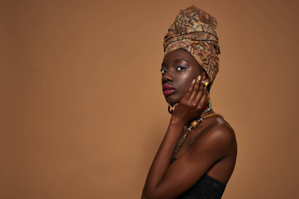 Side view of fashionable black girl wearing traditional african outfit and accessories looking at camera. Pretty young slim woman. Isolated on orange background. Studio shoot. Copy space