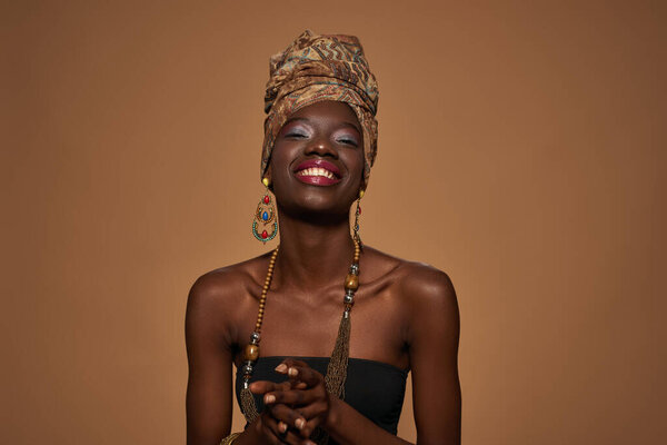 Smiling fashionable black girl wearing african outfit and accessories looking at camera. Young woman wear tank top, turban, necklace, earrings and bracelet. Orange background. Studio shoot. Copy space