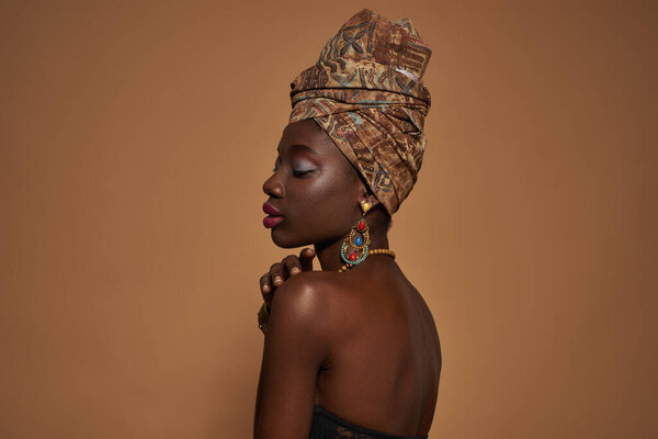 Side view of dreamy stylish black girl with closed eyes wearing traditional african outfit and accessories. Attractive young slim woman. Isolated on orange background. Studio shoot. Copy space