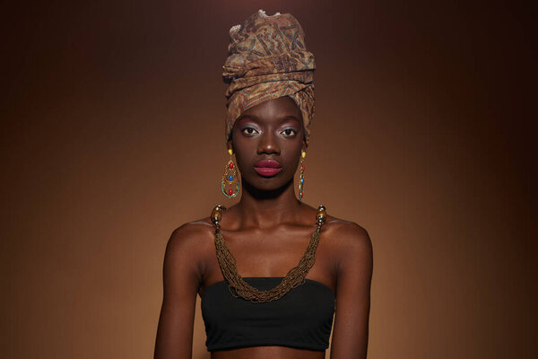 Serious fashionable black girl wearing traditional african outfit and accessories looking at camera. Pretty young slim woman. Isolated on dark orange background. Studio shoot. Copy space