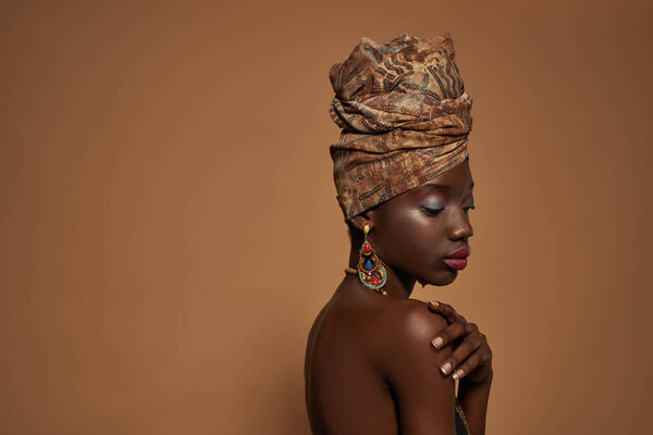 Side view of thoughtful black girl wearing traditional african outfit and accessories. Attractive young woman wear turban, necklace, earrings and bracelet. Orange background. Studio shoot. Copy space