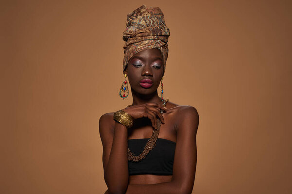 Front view of thoughtful elegant black girl wearing traditional african outfit and accessories. Beautiful young slim woman. Isolated on orange background. Studio shoot. Copy space