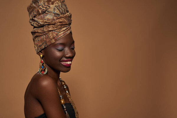 Side view of smiling elegant black girl wearing traditional african outfit and accessories. Young woman wearing tank top, turban, necklace and earrings. Orange background. Studio shoot. Copy space