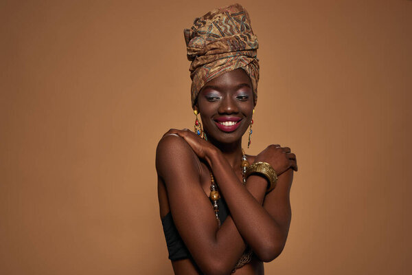Smiling black girl wearing traditional african outfit and accessories hugging herself. Young woman wear tank top, turban, necklace, earrings and bracelet. Orange background. Studio shoot. Copy space