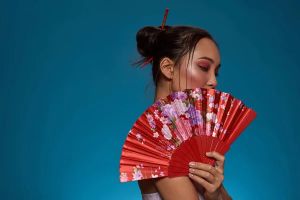 Side view of fashionable asian girl covering part of face with traditional handheld fan. Pretty young brunette slim woman wearing tank top. Female beauty. Blue background. Studio shoot. Copy space