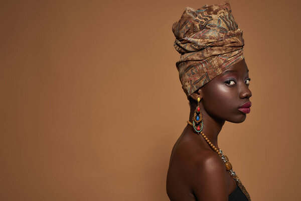 Side view of fashionable black girl wearing african outfit and accessories looking at camera. Young woman wearing tank top, turban, necklace and earrings. Orange background. Studio shoot. Copy space