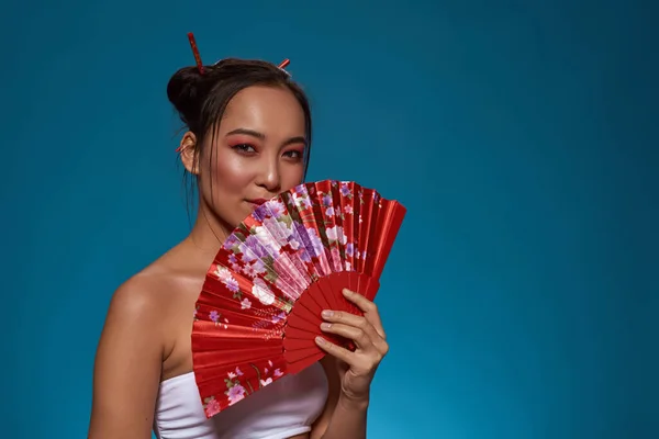 Confident elegant asian girl posing with traditional handheld fan and looking at camera. Beautiful young brunette slim woman wearing tank top. Female beauty. Blue background. Studio shoot. Copy space