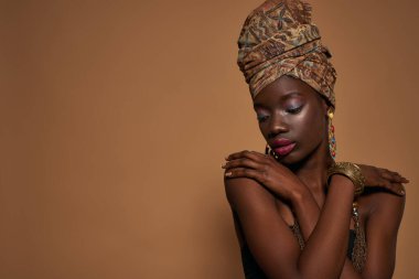 Thoughtful black girl wear traditional african outfit and accessories hug herself. Young woman wearing tank top, turban, necklace, earrings and bracelet. Orange background. Studio shoot. Copy space