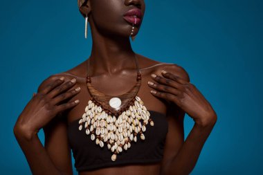 Partial of stylish black girl wearing african outfit and accessories. Attractive young woman wearing tank top, necklace and earrings. Female beauty. Isolated on blue background in studio. Copy space