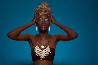 Fashionable black girl looking at camera. Pretty young slim woman wearing tank top and traditional african outfit and accessories. Female beauty. Isolated on blue background. Studio shoot. Copy space
