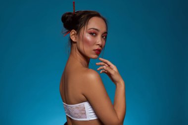 Partial side view of serious stylish asian girl looking at camera. Attractive young brunette slim woman wearing tank top. Female beauty. Isolated on blue background. Studio shoot. Copy space