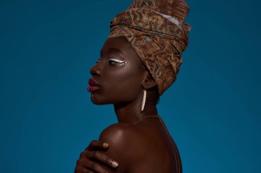 Cropped side view of elegant black girl wearing traditional african outfit and accessories. Beautiful young slim woman wearing turban and earrings. Female beauty. Blue background in studio. Copy space