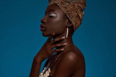 Cropped side view of elegant black girl wearing traditional national african outfit and accessories. Beautiful young slim woman. Female beauty. Isolated on blue background. Studio shoot. Copy space