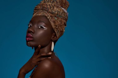 Partial side view of stylish black girl wearing traditional national african outfit and accessories looking at camera. Attractive young woman. Isolated on blue background. Studio shoot. Copy space