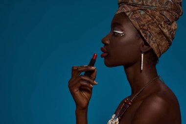 Partial side view of stylish black girl painting lips with lipstick. Attractive young woman wearing traditional african outfit and accessories. Female beauty. Blue background. Studio shoot. Copy space