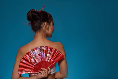 Back view of stylish asian girl with traditional handheld fan. Attractive young brunette slim woman wearing tank top. Female beauty. Isolated on blue background. Studio shoot. Copy space