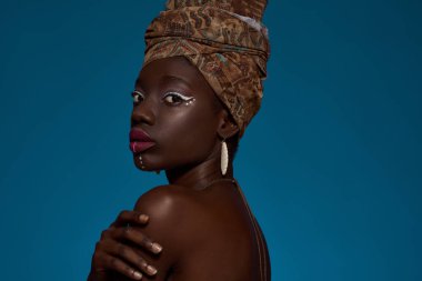 Partial side view of fashionable black girl wearing african outfit and accessories looking at camera. Pretty young woman wear turban and earrings. Isolated on blue background. Studio shoot. Copy space