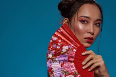 Cropped of serious fashionable asian girl posing with traditional handheld fan and looking at camera. Pretty young brunette woman. Female beauty. Isolated on blue background. Studio shoot. Copy space