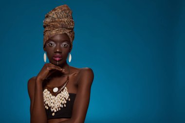 Serious elegant black girl looking at camera. Beautiful young slim woman wearing traditional african outfit and accessories. Female beauty. Isolated on blue background. Studio shoot. Copy space
