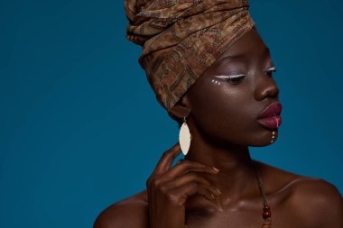Cropped of stylish black girl. Attractive young slim woman wearing wearing traditional national african turban and earrings. Female beauty. Isolated on blue background. Studio shoot. Copy space