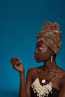 Profile of focused stylish black girl looking away. Attractive young slim woman wearing traditional african outfit and accessories. Female beauty. Isolated on blue background. Studio shoot. Copy space