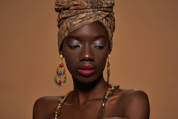 Cropped of stylish black girl wearing traditional african outfit and accessories. Attractive young slim woman wearing turban, necklace and earrings. Isolated on orange background in studio. Copy space