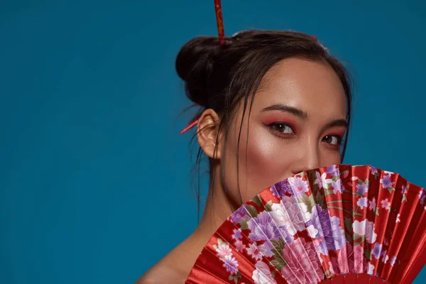 Partial of elegant asian girl covering part of face with traditional handheld fan. Beautiful young brunette woman look at camera. Female beauty. Isolated on blue background. Studio shoot. Copy space