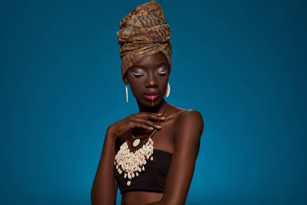 Thoughtful elegant black girl wearing african outfit and accessories. Beautiful young woman wearing tank top, turban, necklace and earrings. Female beauty. Blue background. Studio shoot. Copy space