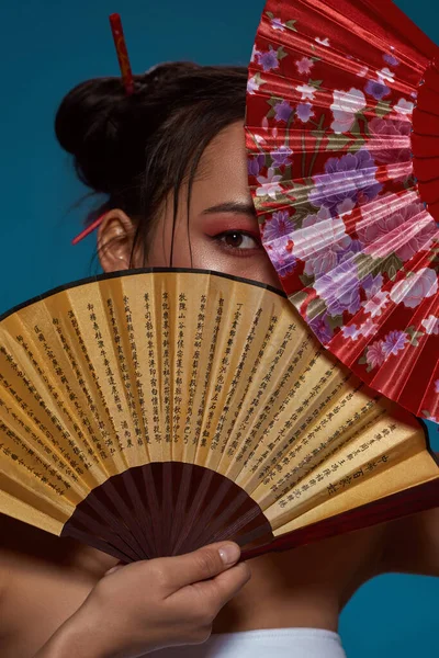 Stylish asian girl covering face with two traditional handheld fans. Attractive young brunette slim woman wearing tank top looking at camera. Female beauty. Isolated on blue background. Studio shoot