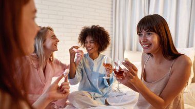 Young joyful multiracial girlfriends drink wine and feed marshmallow each other on bed during girlish sleepover at night home. Black and european zoomer girls. Friendship. Entertainment and leisure
