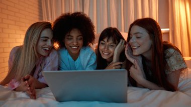 Young smiling multiethnic girlfriends watching laptop on bed during girlish sleepover at night. Black and caucasian zoomer girls rest and enjoy time together. Friendship. Entertainment and leisure