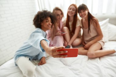 Focus on foreground on smartphone during blurred multiethnic girlfriends taking selfie on bed at girlish pajama party at home. Black and caucasian zoomer girls. Friendship. Entertainment and leisure