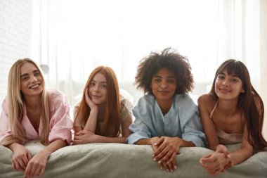 Group of young smiling multiethnic girlfriends lying on bed and looking at camera during girlish pajama party. Black and caucasian zoomer girls resting and spending time together. Friendship. Bedroom