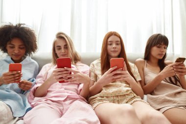 Young multiethnic girlfriends typing and chatting smartphones on bed during girlish pajama party at home. Black and caucasian zoomer girls resting together. Friendship. Entertainment and leisure