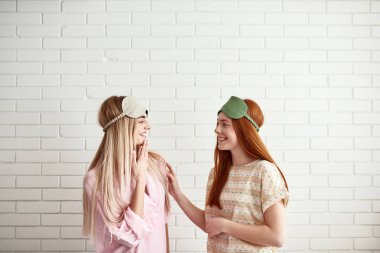 Side view of young caucasian smiling girls looking at each other on wall background at home. Teen female zoomers wearing homewear and sleep masks resting and spending time together. Friendship