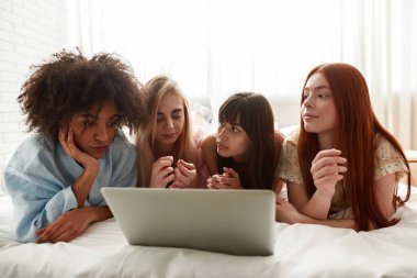 Young multiracial girlfriends lying and resting on bed during watching laptop at girlish pajama party. Black and caucasian zoomer girls spending time together. Friendship. Entertainment and leisure