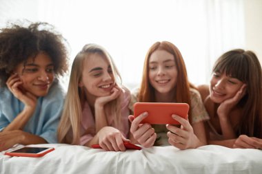 Selective focus on smartphone in hand of red haired girl show something to multiethnic girlfriends on bed during girlish pajama party. Young smiling girls. Friendship. Rest, entertainment and leisure