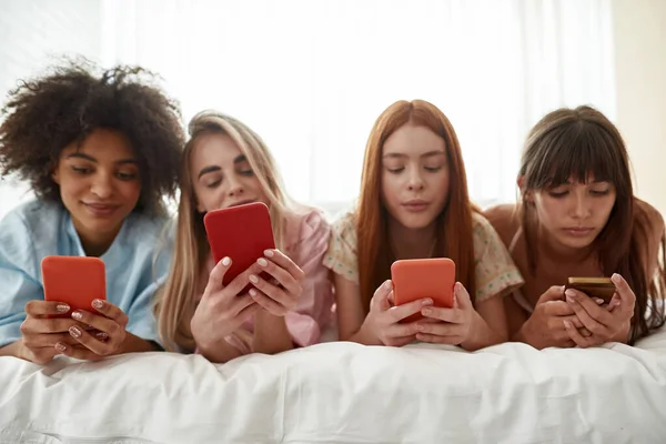 Young multiethnic girlfriends in row using smartphones on bed during girlish pajama party at home. Black and caucasian zoomer girls rest and spend time together. Friendship. Entertainment and leisure