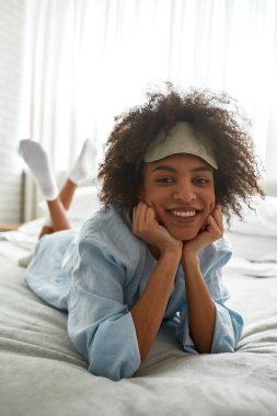 Young smiling black girl lying, resting and looking at camera on bed at home in morning. Curly teen female zoomer wearing homewear and sleep mask. Domestic lifestyle. Modern bedroom in spacious flat