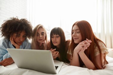 Young multiracial girlfriends watching laptop on bed during girlish pajama party at home. Black and caucasian zoomer girls resting and enjoying time together. Friendship. Entertainment and leisure